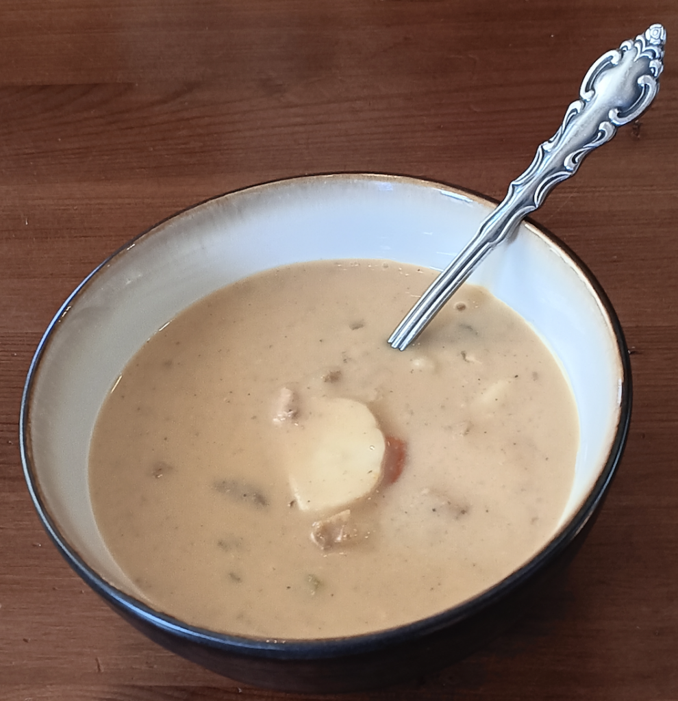 a bowl of soup with a slice of potato as garnish; a silver spoon sits inside the bowl.